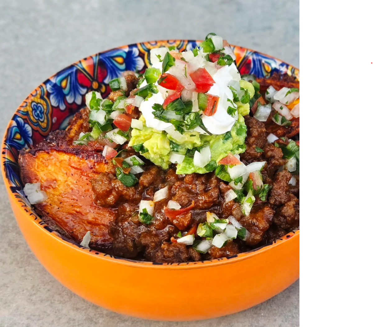 A colorful bowl filled with chili, topped with guacamole, sour cream, and fresh salsa, sits on a gray surface. Fried plantains are visible along the edge of the bowl.