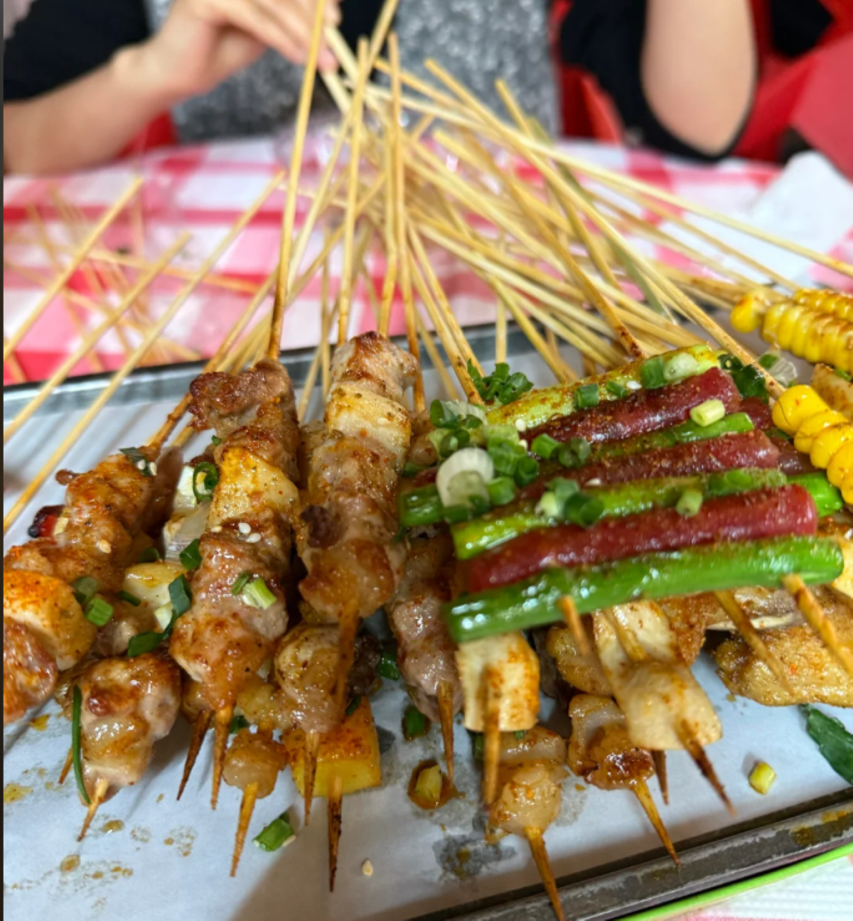Assorted grilled skewers with meat, green beans, sausage, and corn, seasoned with spices and chopped scallions, served on a tray. A person sits in the background holding skewers.
