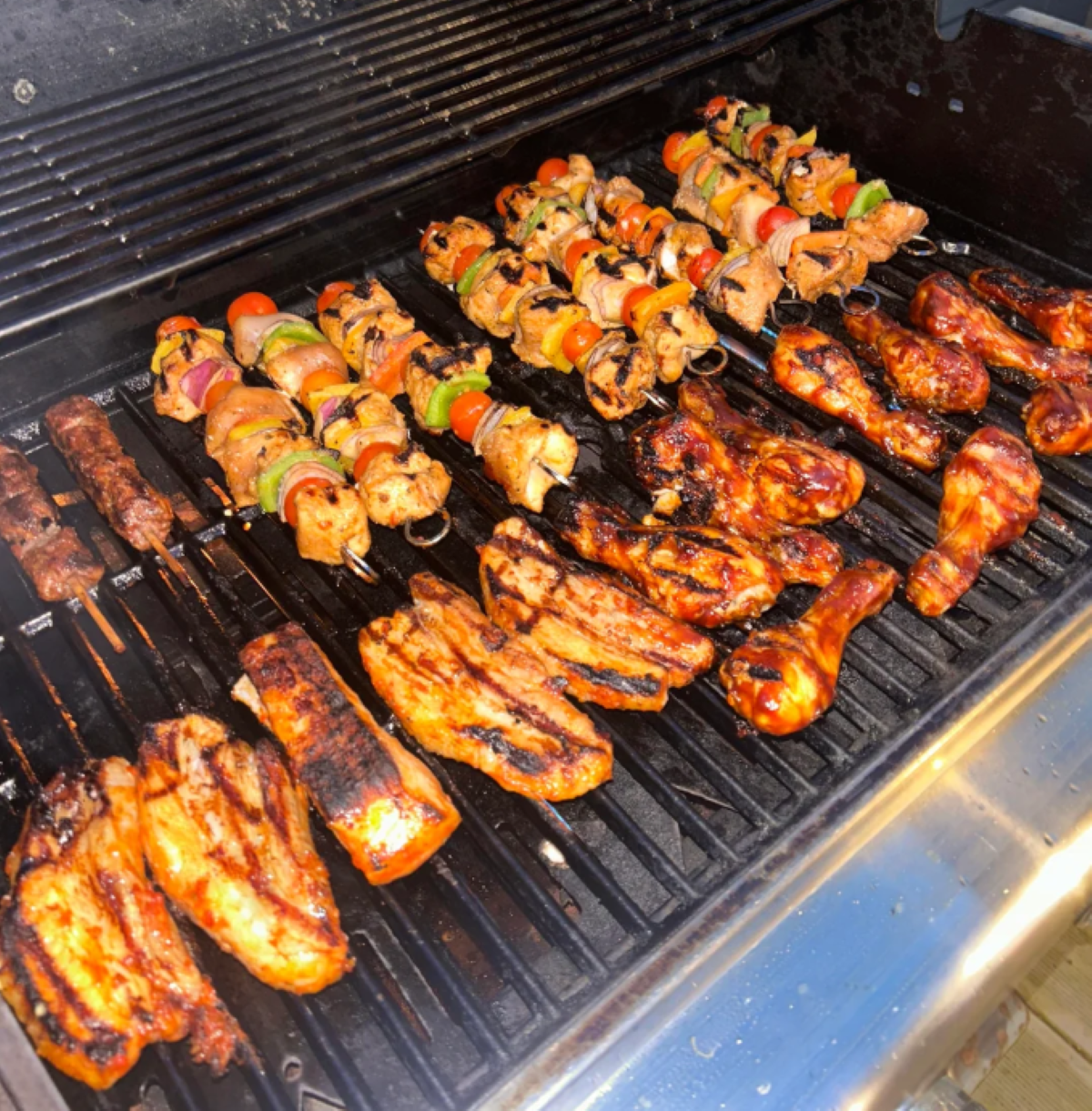 A grill loaded with skewered kebabs, marinated chicken drumsticks, and grilled chicken breasts—all with visible grill marks and colorful vegetables—cooking over open flames, sets the scene for one of the best BBQ cookouts this summer.