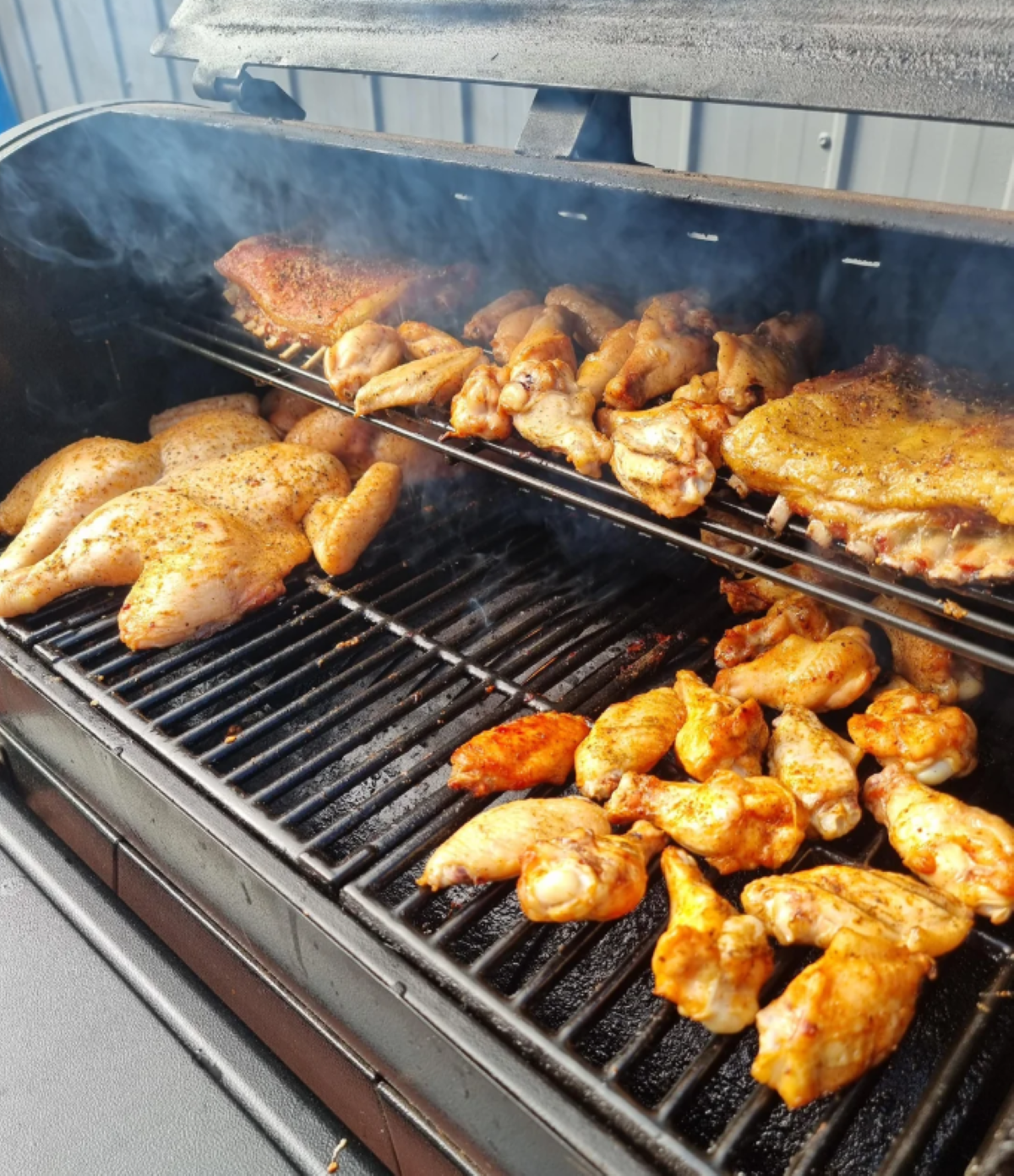 Chicken pieces and wings are being grilled on a smoky outdoor barbecue, with both upper and lower racks filled with seasoned meat—perfect for hosting the best BBQ cookouts this summer.