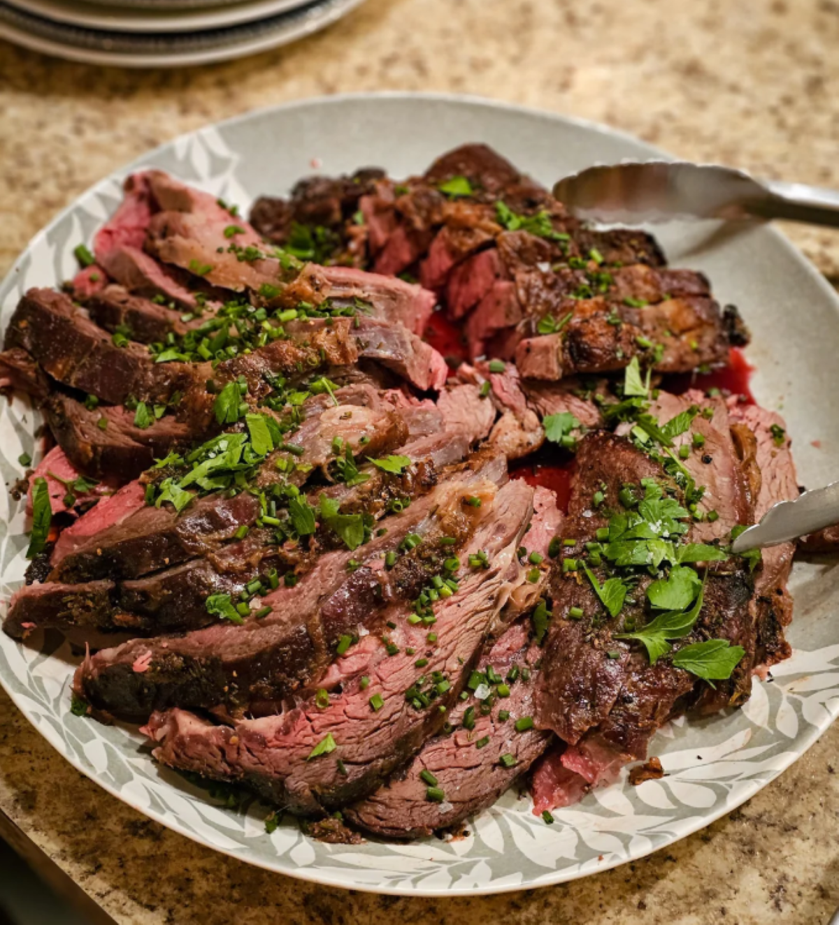 Sliced roast beef topped with chopped fresh herbs, arranged on a round plate with metal tongs on the side, sitting on a speckled countertop.