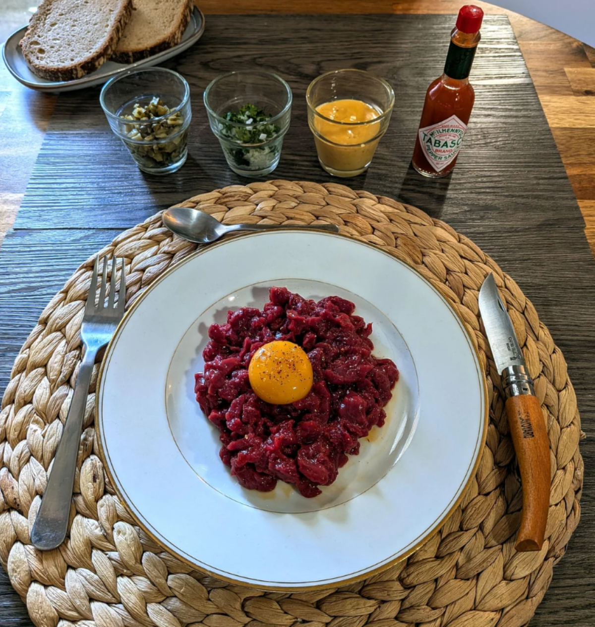A plate of beef tartare topped with an egg yolk, surrounded by bread slices, herbs, sauce, capers, and Tabasco on a woven placemat, with cutlery beside the plate.