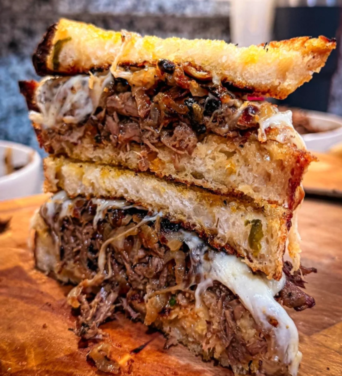 A close-up of a stacked grilled cheese sandwich filled with shredded beef, caramelized onions, and melted cheese, sitting on a wooden cutting board.