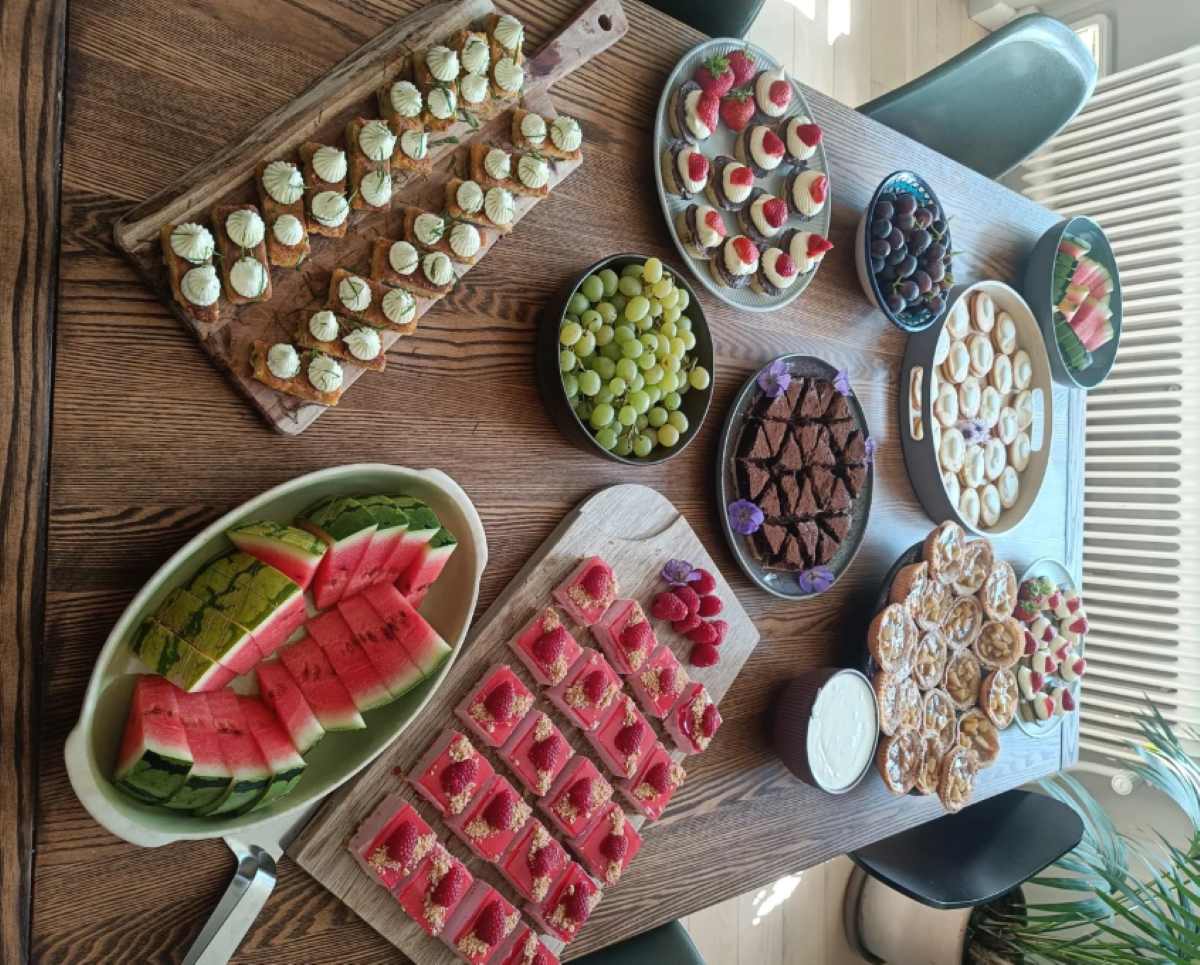 A wooden table set with watermelon slices, grapes, brownies, cookies, tarts, and an assortment of finger foods arranged on boards and plates. Natural light streams in from a nearby window.
