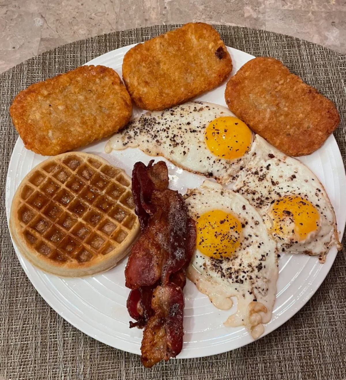 A plate with two sunny-side-up eggs, three crispy hash browns, three strips of bacon, and a waffle with syrup, on a textured placemat.