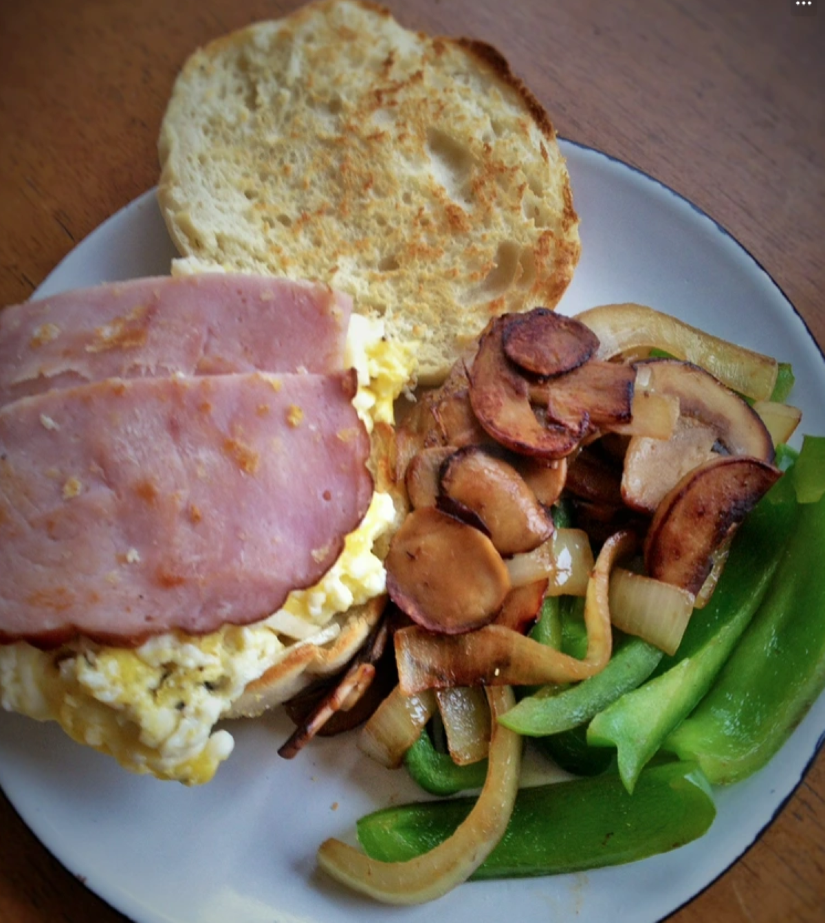 Breakfast sandwich with scrambled eggs and ham on an English muffin, served with sautéed mushrooms, onions, and sliced green bell peppers on a white plate.