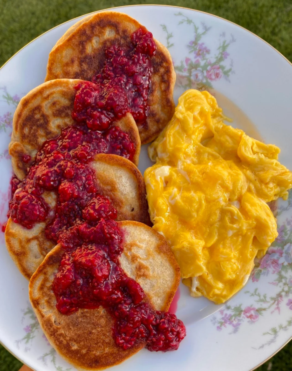 A plate with three pancakes topped with raspberry sauce, served alongside a portion of scrambled eggs. The plate has a floral pattern and sits on a green grassy surface.