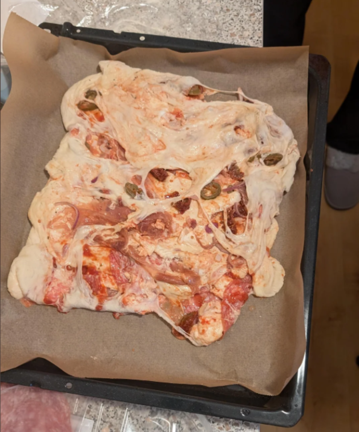 A misshapen homemade pizza with unevenly melted cheese, visible jalapeño slices, sauce, and dough on parchment paper atop a baking tray—one of those hilarious cooking disasters that tastes far better than it looks.
