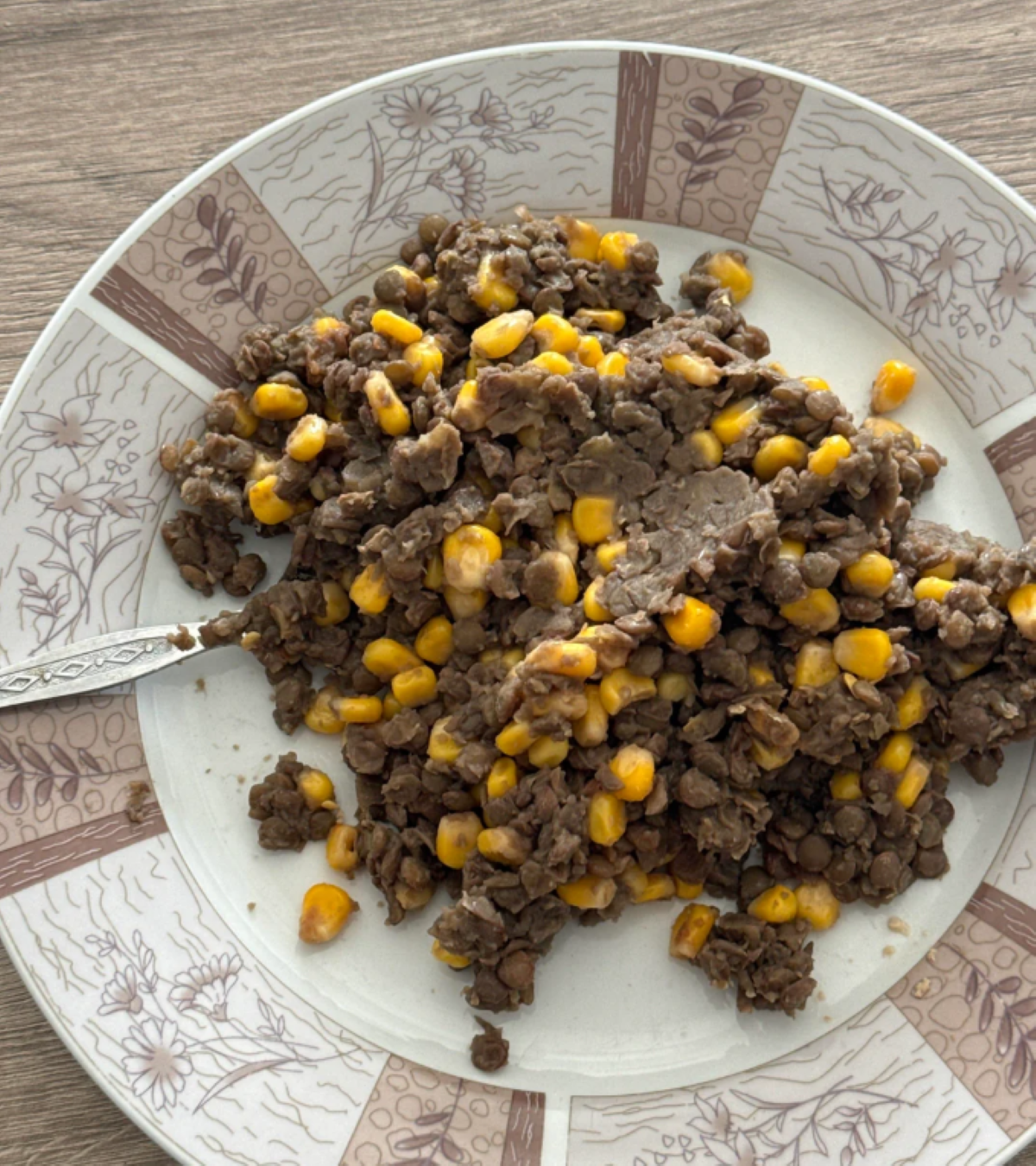 A plate with a mixture of cooked lentils and yellow corn, served with a spoon, on a patterned plate placed on a wooden surface—thankfully not the result of one of my hilarious cooking disasters!