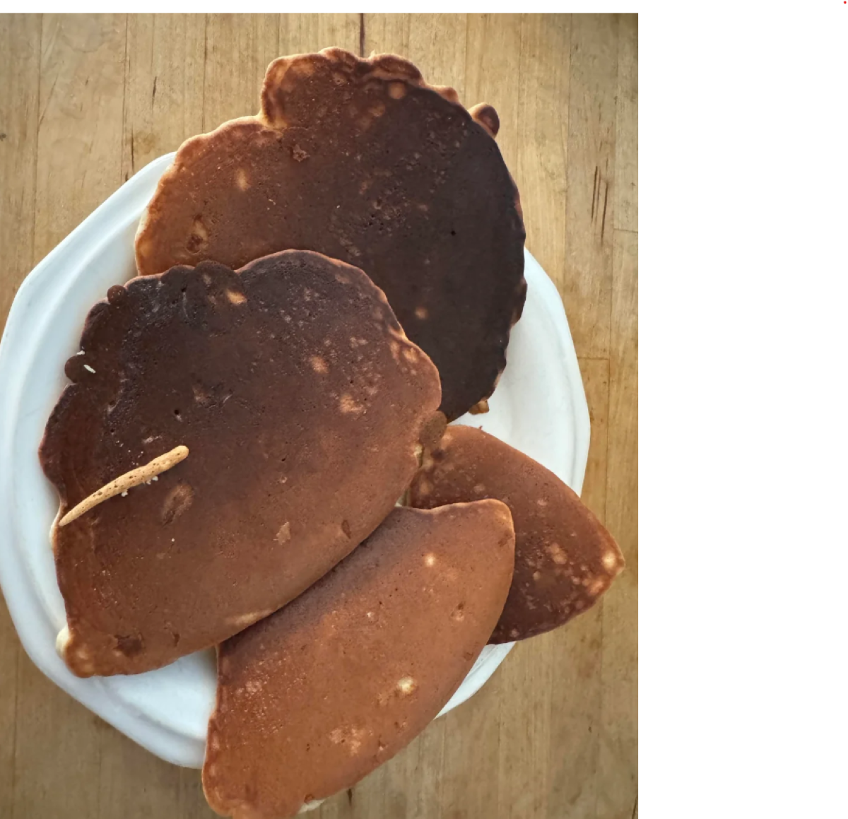 Four unevenly cooked pancakes, two dark and two lighter, stacked on a white plate placed on a wooden surface.