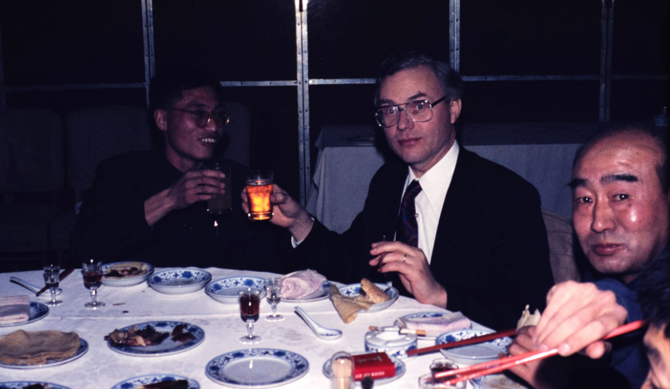 Three men sit around a table with plates of food, drinks, and chopsticks. Two of them are raising glasses, appearing to make a toast, while the third uses chopsticks. The dimly lit, formal setting evokes the warmth and camaraderie of nostalgic dinners shared among friends.