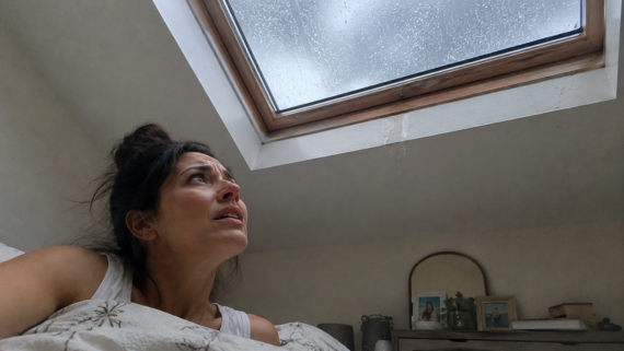 A woman in bed looks worriedly at a skylight window with rain pouring down, concerned about possible water leakage&mdash;a scenario that highlights why skylights are now among the home features homeowners no longer want.