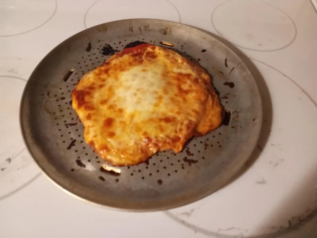 A misshapen, baked cheese pizza sits on a round metal pizza pan atop a white stovetop. The pizza appears slightly overcooked and uneven.