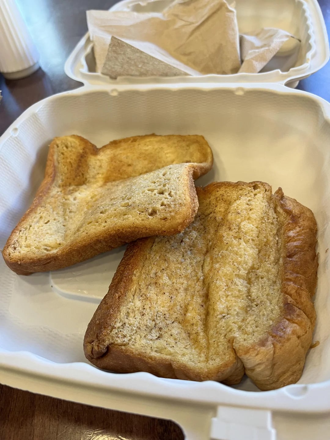 Two pieces of toasted bread with uneven shapes sit in a white takeout container. A napkin is visible in the background.