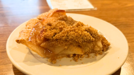 A slice of apple pie with a crumb topping sits on a white plate on a wooden table, evoking the timeless comfort of homemade apple pies, with a blurred receipt or paper in the background.