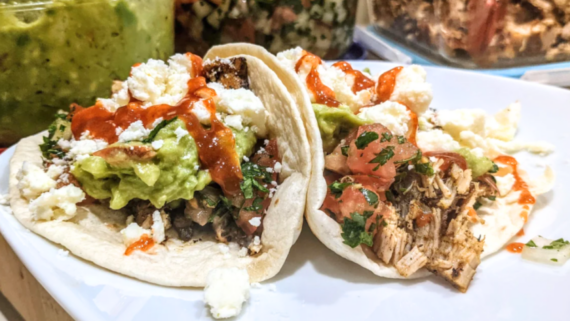 Two soft tacos filled with shredded meat, guacamole, crumbled cheese, chopped herbs, and salsa sit on a white plate&mdash;an inviting glimpse of delicious meals to come. Containers of guacamole and other toppings are blurred in the background.
