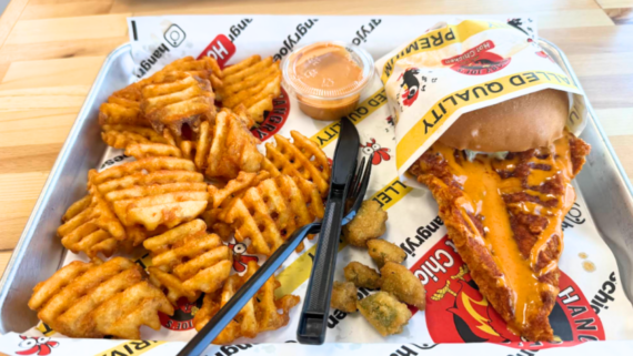 A tray with waffle fries, fried okra, and a fried chicken sandwich drizzled with orange sauce&mdash;a standout item from one of the fastest-growing fried chicken chains&mdash;served alongside a cup of dipping sauce, plus a black plastic fork and knife on branded paper.