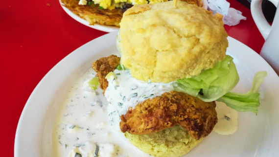 A fried chicken biscuit sandwich with lettuce and creamy white sauce sits on a white plate, showcasing why it ranks among the best chicken sandwiches, with part of another breakfast dish in the background.
