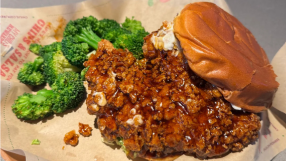 A crispy fried chicken sandwich with sauce is served on a bun, accompanied by a side of steamed broccoli on a brown paper tray&mdash;perfect for fast-food menu tests seeking tasty new options.