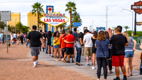 A long line of people, perhaps making one of the classic tourist mistakes, waits near the "Welcome to Fabulous Las Vegas, Nevada" sign on a sunny day, with palm trees and Las Vegas hotels visible in the background.
