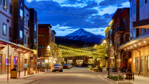 A cozy mountain town street at dusk, reminiscent of the charm found in some of the world&rsquo;s top 20 travel destinations, is softly illuminated by string lights overhead. Cars are parked along the road while inviting shops and charming buildings line each side, all set against a breathtaking backdrop&mdash;a snow-capped mountain rising beneath a blue, cloud-streaked sky.