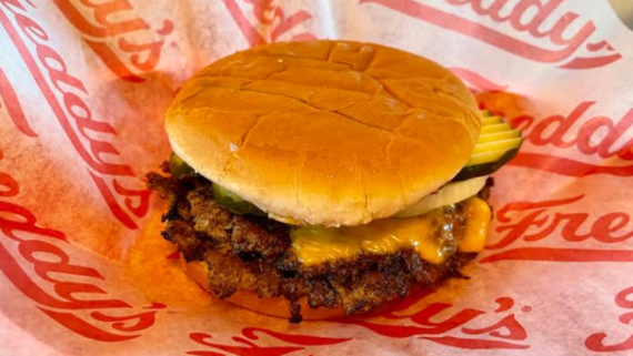 A classic fast food burger, this cheeseburger features pickles and melted cheese on a toasted bun, served on red and white Freddy&rsquo;s branded paper.