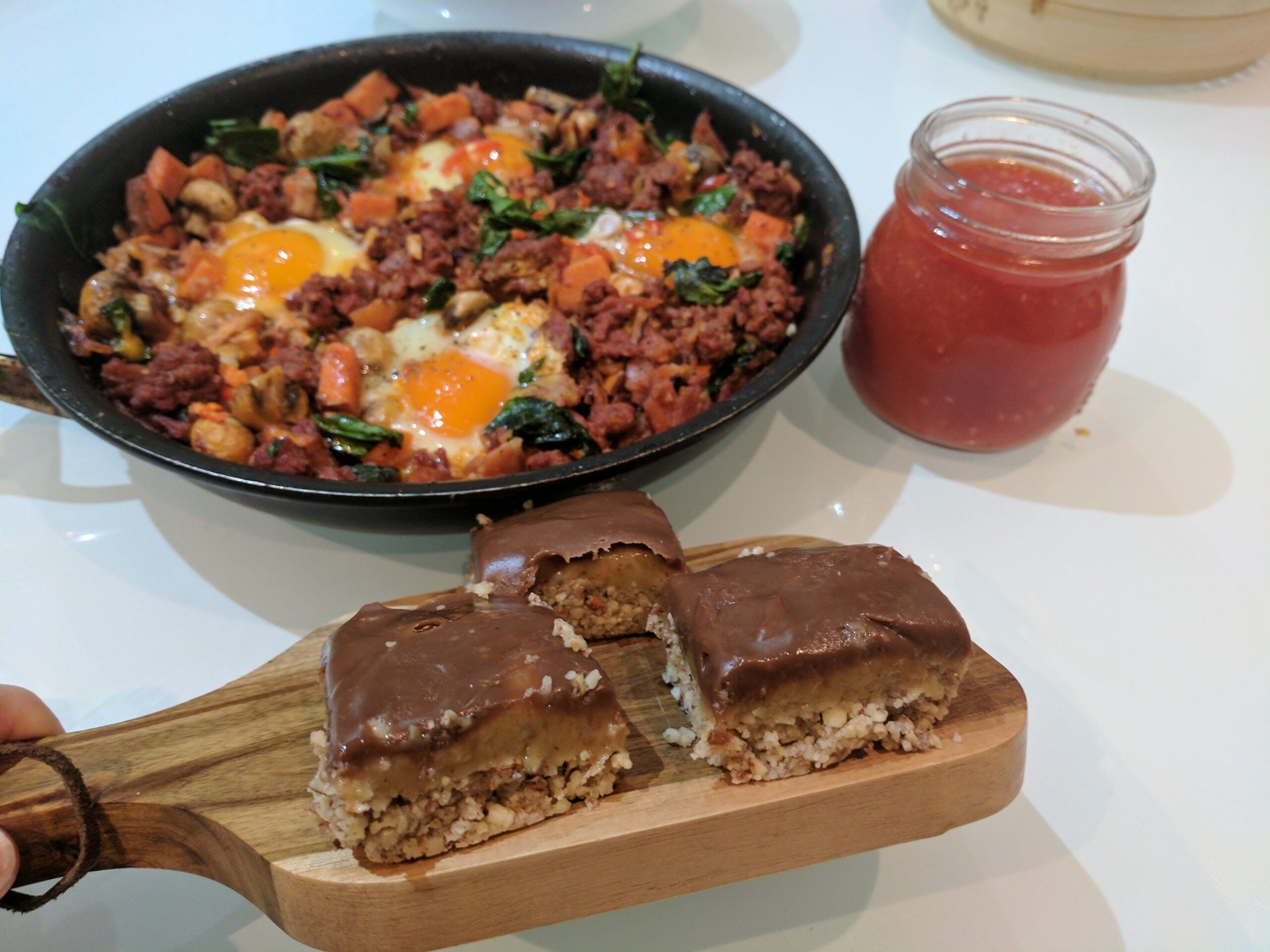 A skillet with eggs, vegetables, and ground meat, a jar of tomato sauce, and three chocolate-covered dessert bars on a wooden board are displayed on a white table.