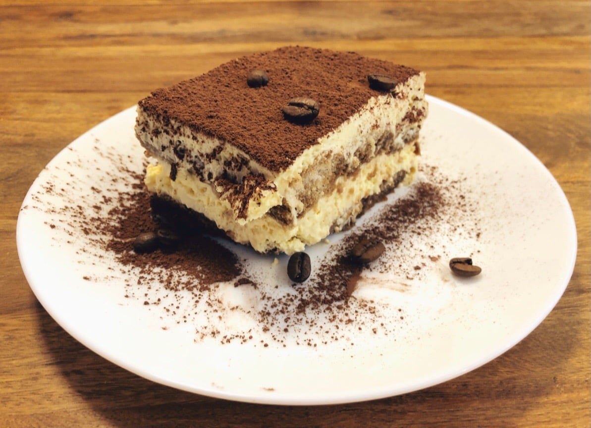 A slice of tiramisu topped with cocoa powder and coffee beans sits on a white plate, with cocoa powder sprinkled around it, on a wooden table—a perfect treat for lovers of homemade desserts.