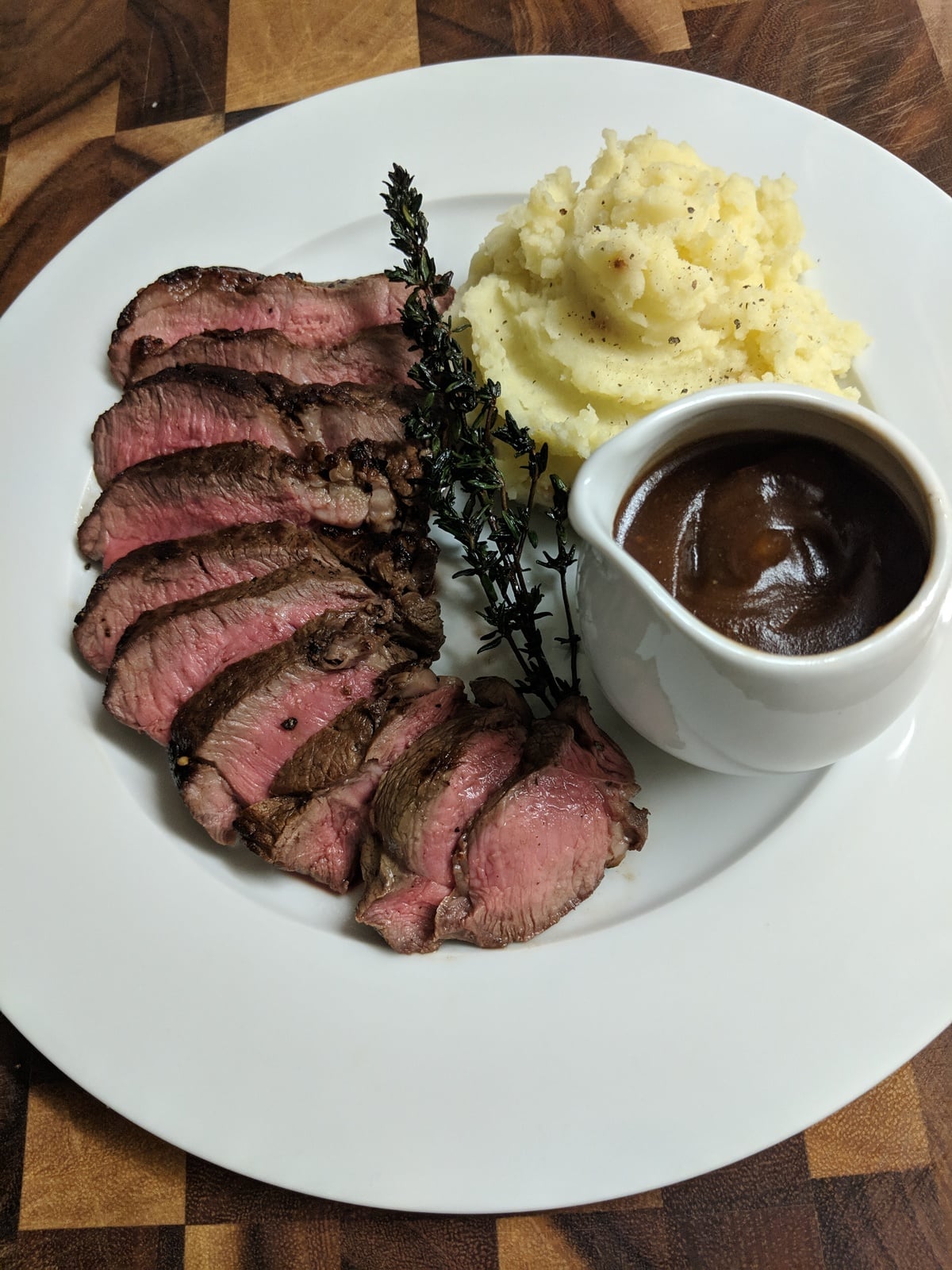 Sliced medium-rare steak with sprig of thyme, mashed potatoes, and a cup of brown gravy on a white plate, placed on a wooden surface.