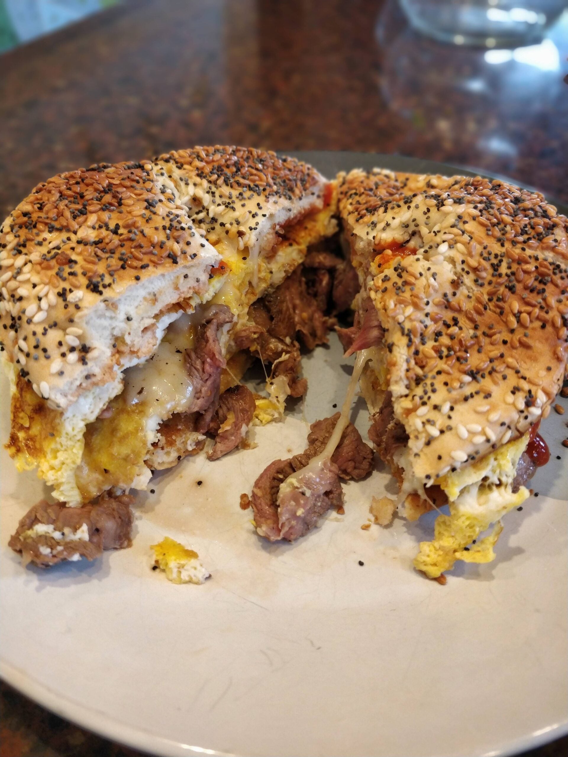 A sesame seed bagel sandwich cut in half, filled with scrambled eggs, melted cheese, and pieces of cooked meat, served on a white plate.