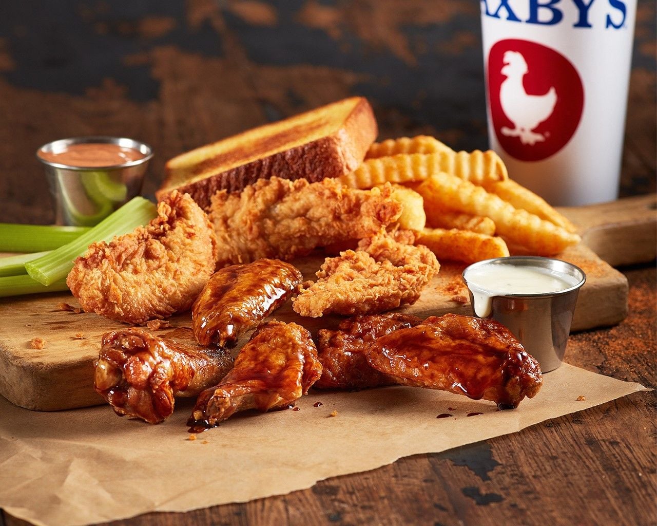 A meal with fried chicken tenders, sauced chicken wings, crinkle-cut fries, celery sticks, dipping sauces, toast, and a drink in a Zaxby’s cup—all served on a wooden board with paper underneath—perfectly showcases why Zaxby’s stands out among fried chicken chains.