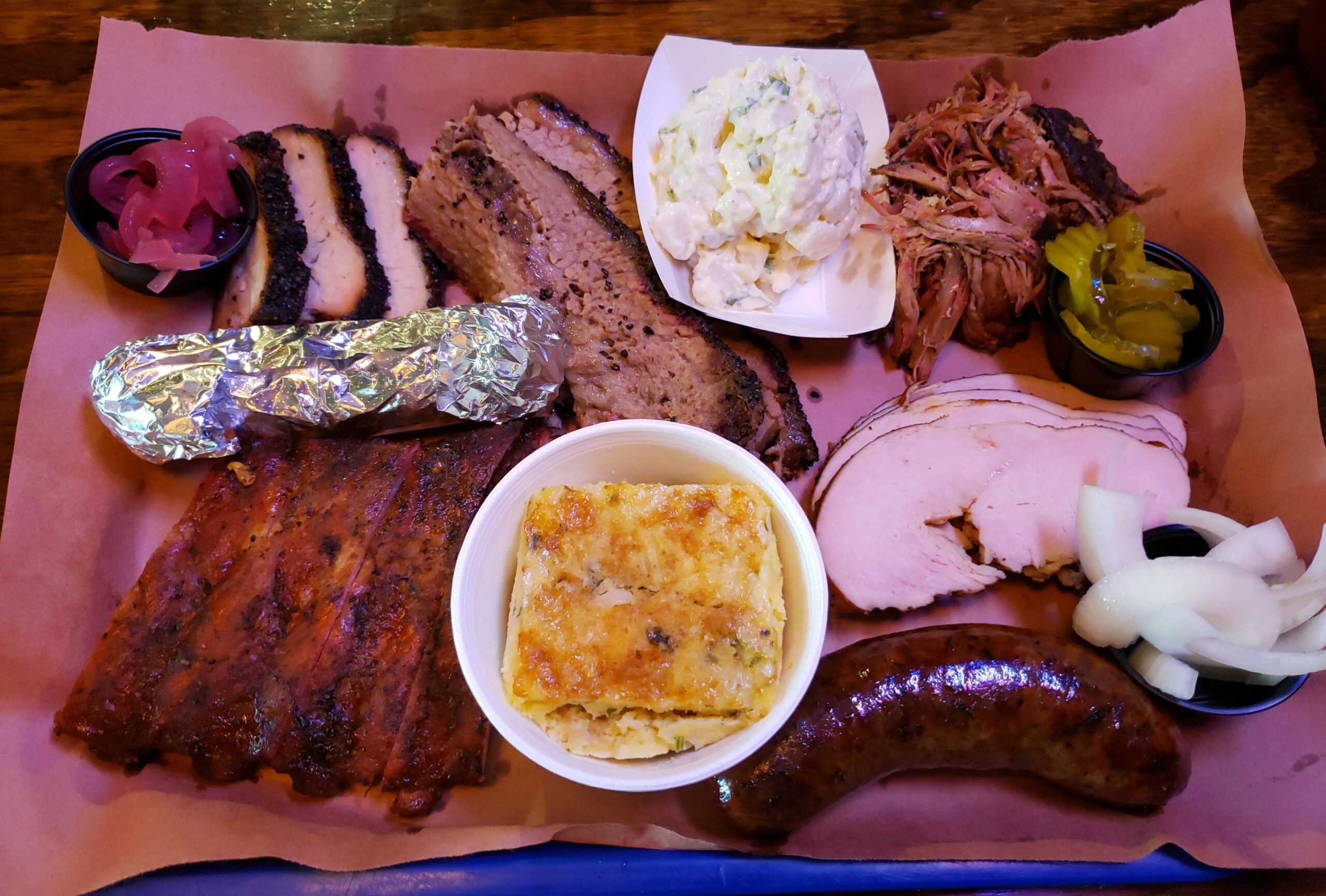 A tray of barbecue with sliced brisket, pork, turkey, smoked sausage, ribs, potato salad, cornbread, pickles, onions, and a foil-wrapped baked potato on brown paper—perfect for the best BBQ cookouts this summer.