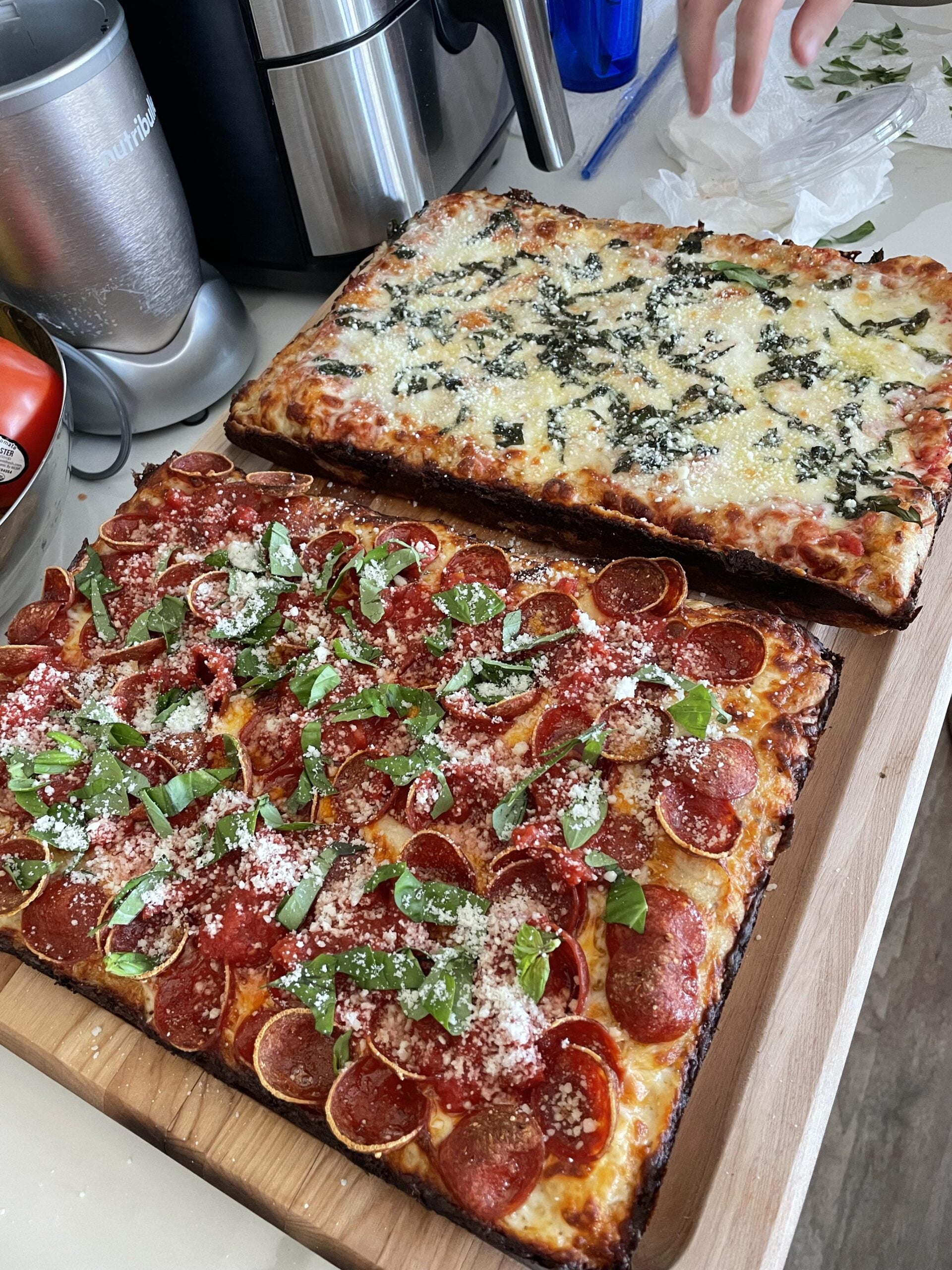 Two rectangular pizzas on a wooden board: one topped with pepperoni, basil, and cheese, the other with cheese and greens. Kitchen appliances and a hand appear in the background.