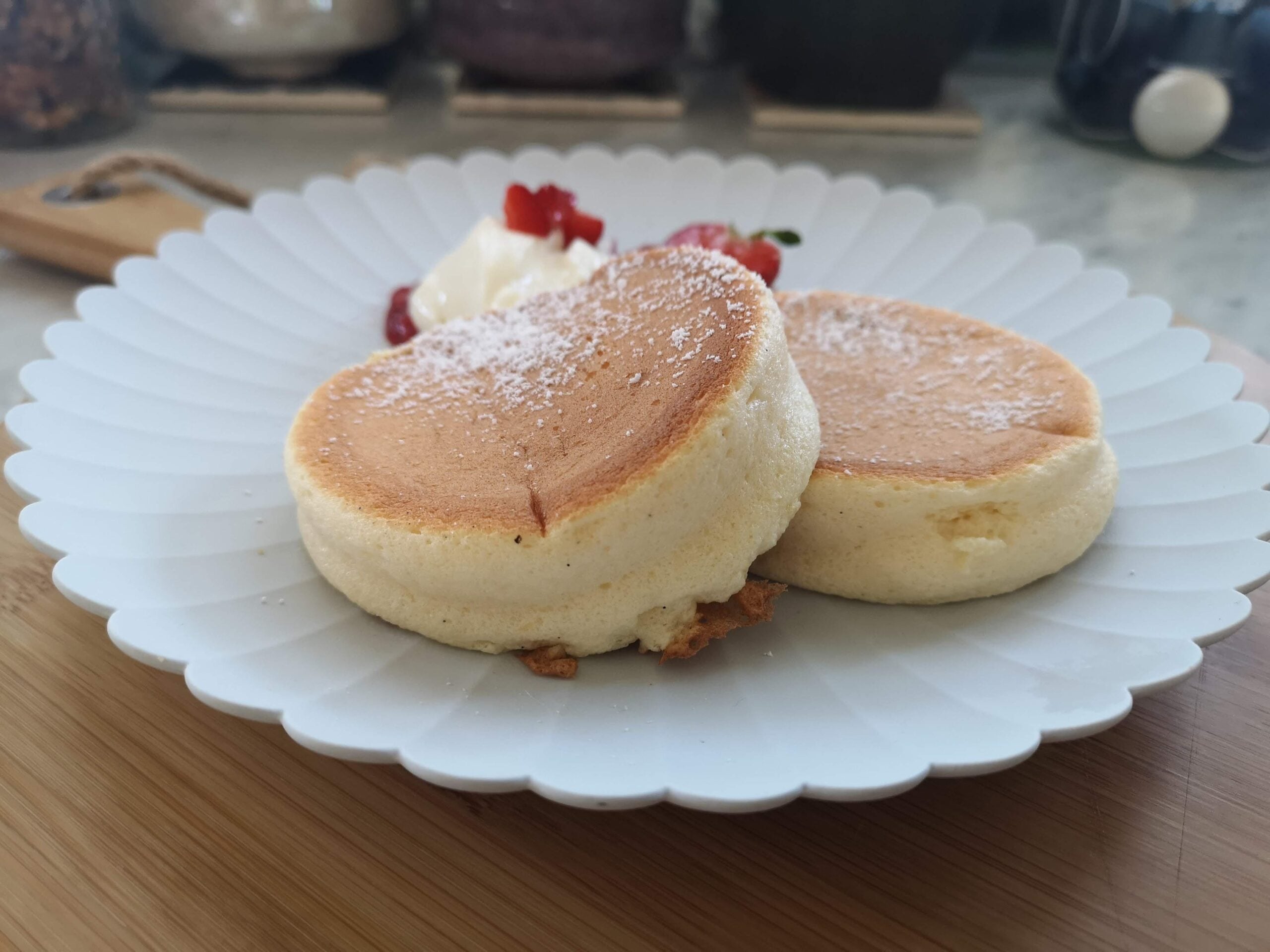 Two fluffy homemade pancakes dusted with powdered sugar sit on a white plate, served with whipped cream and strawberry slices in the background.