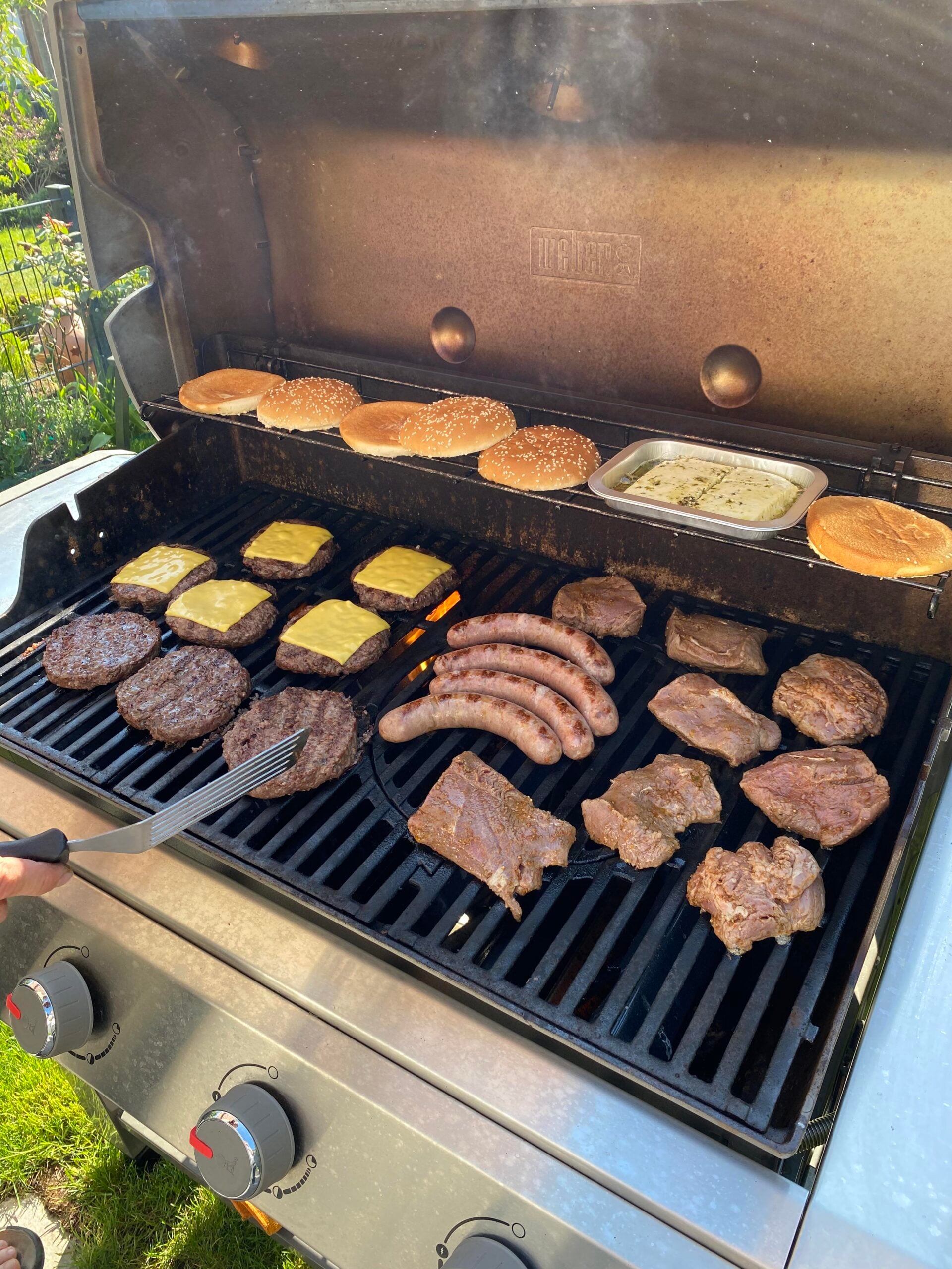 An outdoor grill filled with burger patties topped with cheese, sausages, steaks, buns, and a tray of sliced onions. A spatula is flipping food while sunlight brightens the scene—capturing the essence of the best BBQ cookouts this summer.