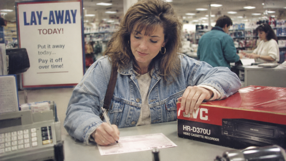 A woman in a denim jacket fills out a form at a store counter with a JVC VCR beside her, taking advantage of the store features. A &ldquo;LAY-AWAY TODAY!&rdquo; sign and other shoppers are visible in the background.