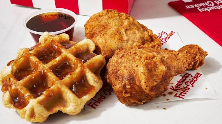 A serving of fried chicken drumstick and thigh with a waffle, topped with syrup, on a Kentucky Fried Chicken wrapper. A small cup of syrup is in the background.