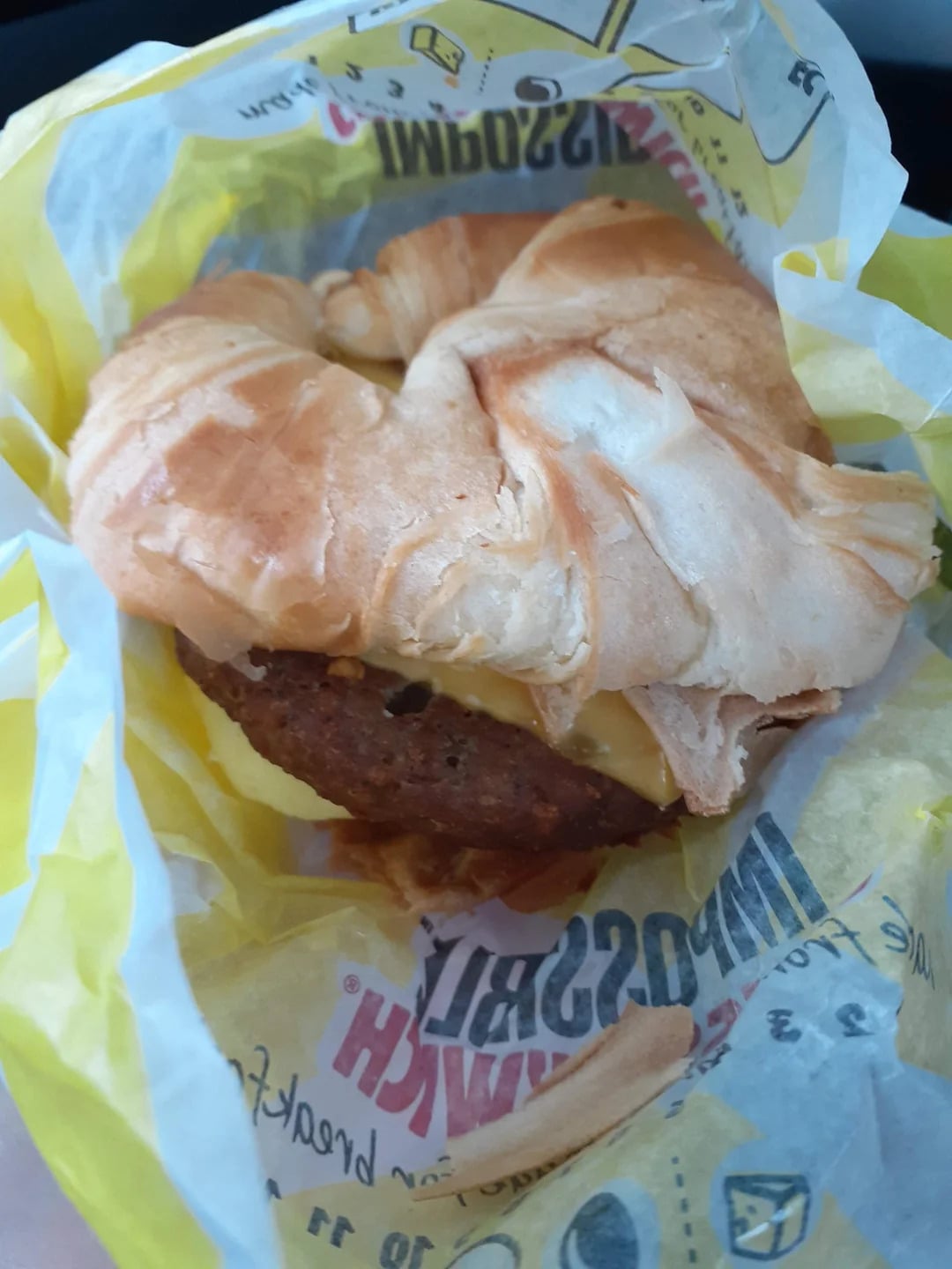 A croissant breakfast sandwich with a sausage patty and melted cheese, partially wrapped in yellow and white branded paper.