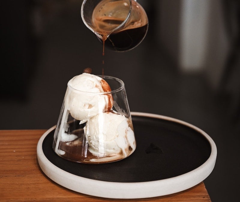 A hand pours espresso from a small glass pitcher over two scoops of vanilla ice cream in a clear glass, creating an affogato dessert—a treat often found in cozy coffee houses. The glass sits on a round black tray atop a wooden table.