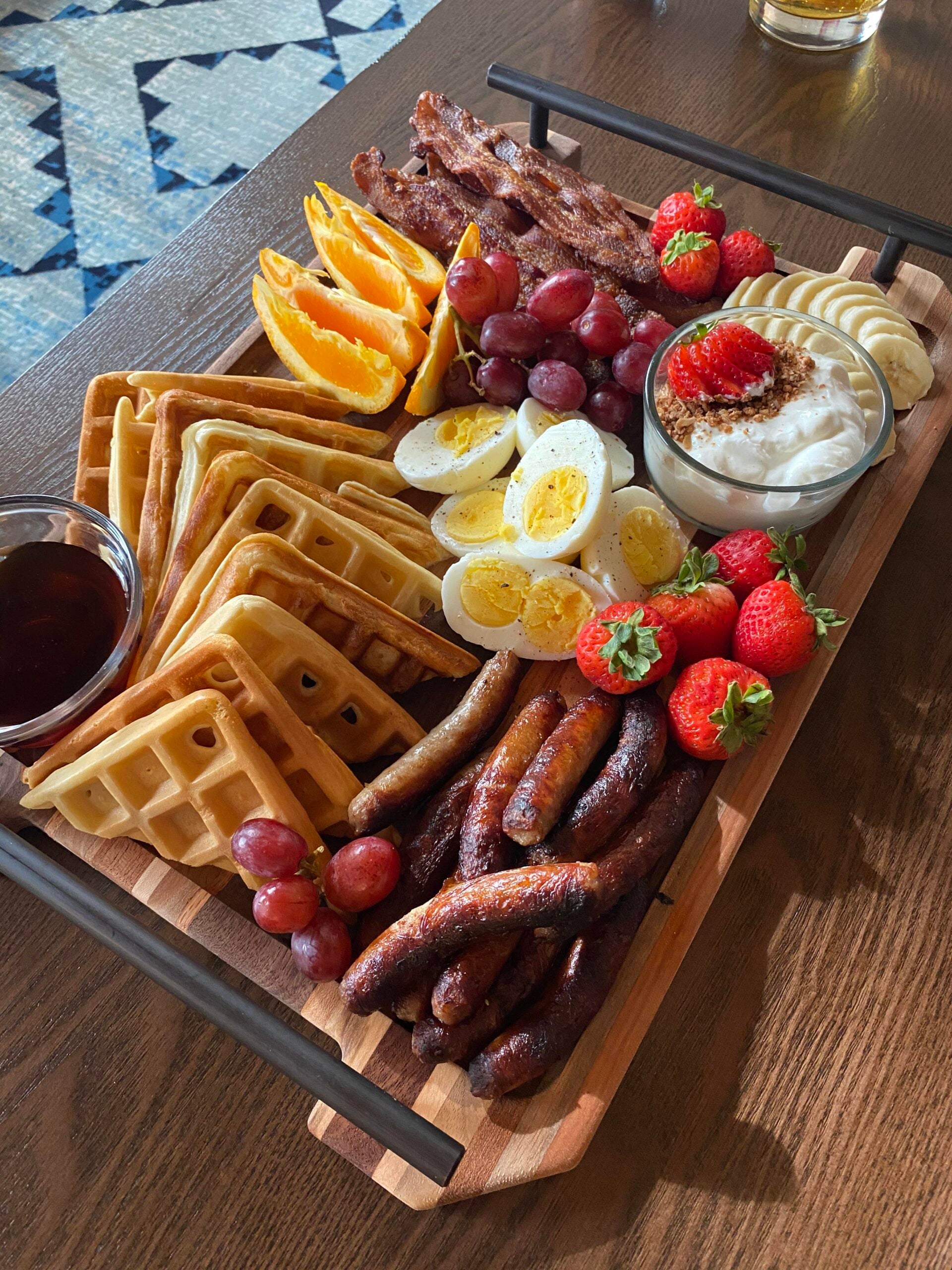 A wooden tray with waffles, syrup, bacon, sausage, hard-boiled eggs, grapes, orange slices, strawberries, banana slices, and a bowl of yogurt topped with cinnamon, set on a table.