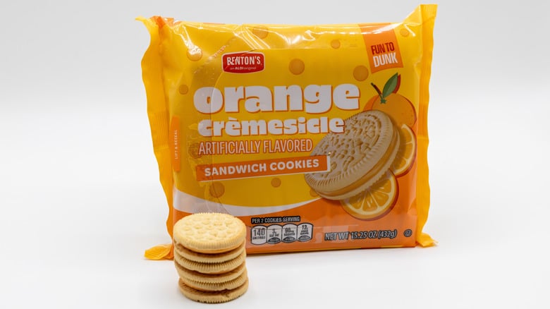 A package of Benton's Orange Crèmesicle sandwich cookies sits behind a stack of five cookies, with orange slices and a cream-filled cookie pictured on the packaging.