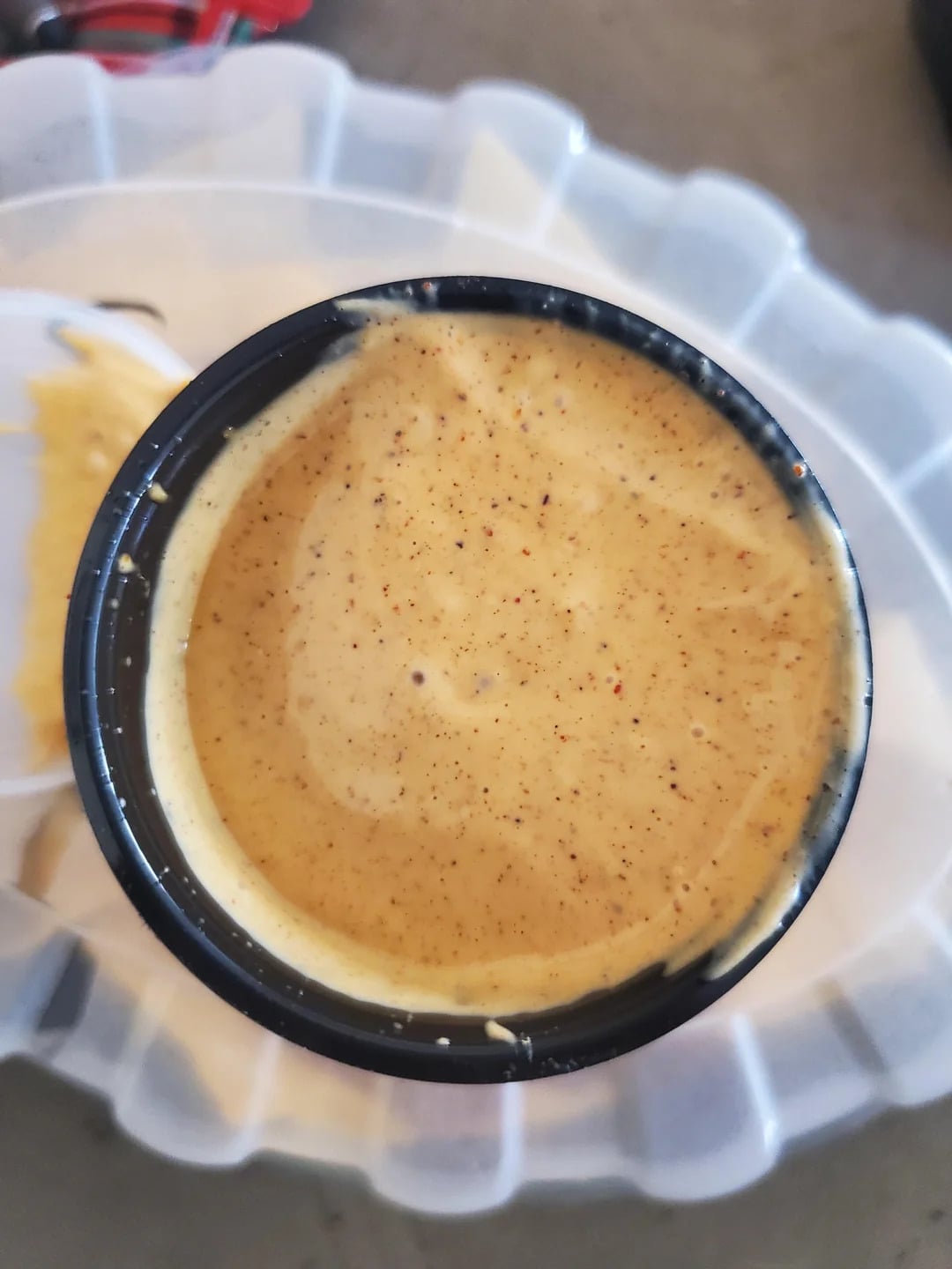 Close-up of a small black plastic cup filled with creamy, light orange dipping sauce, speckled with visible seasoning, placed on a translucent plastic tray—reminiscent of the flavorful condiments often found alongside popular fast-food grocery store products.