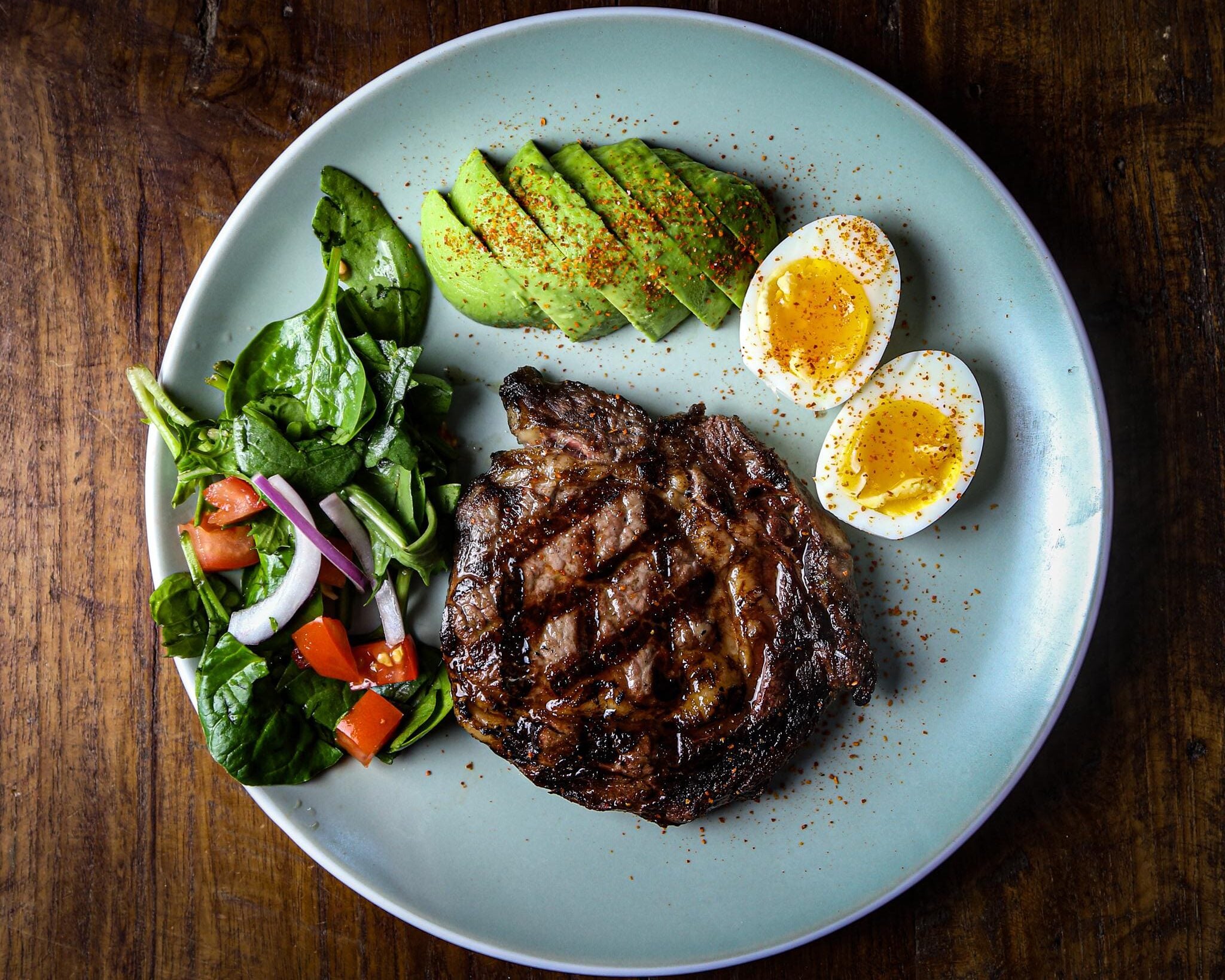 A plate with grilled steak, sliced avocado, two halved soft-boiled eggs sprinkled with seasoning, and a fresh salad with spinach, tomato, and red onion on a wooden table.