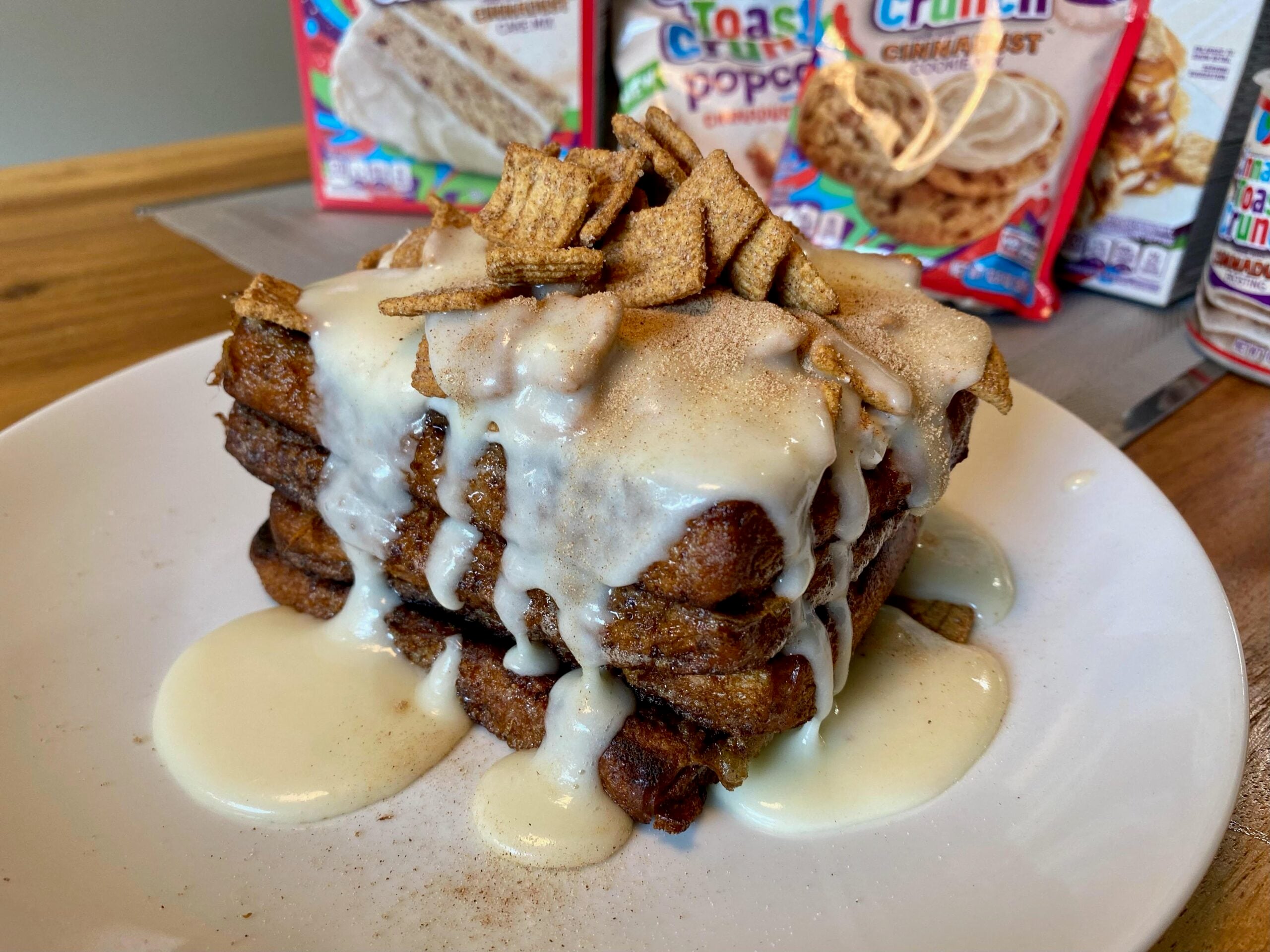 A stack of French toast topped with white icing and Toast Crunch cereal pieces, with cereal snack bags in the background.