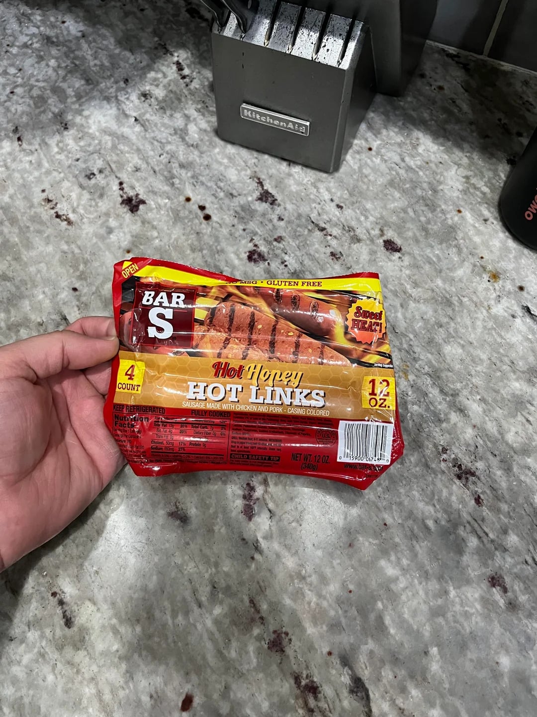 A hand holds a package of Bar-S Hot Honey Hot Links with four sausages, placed on a gray marble countertop near a knife block.