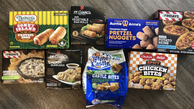 Assorted frozen appetizers including corn dogs, egg rolls, pretzel nuggets, potato skins, chicken dumplings, spinach artichoke dip, castle bites, and spicy chicken bites on a wooden surface.