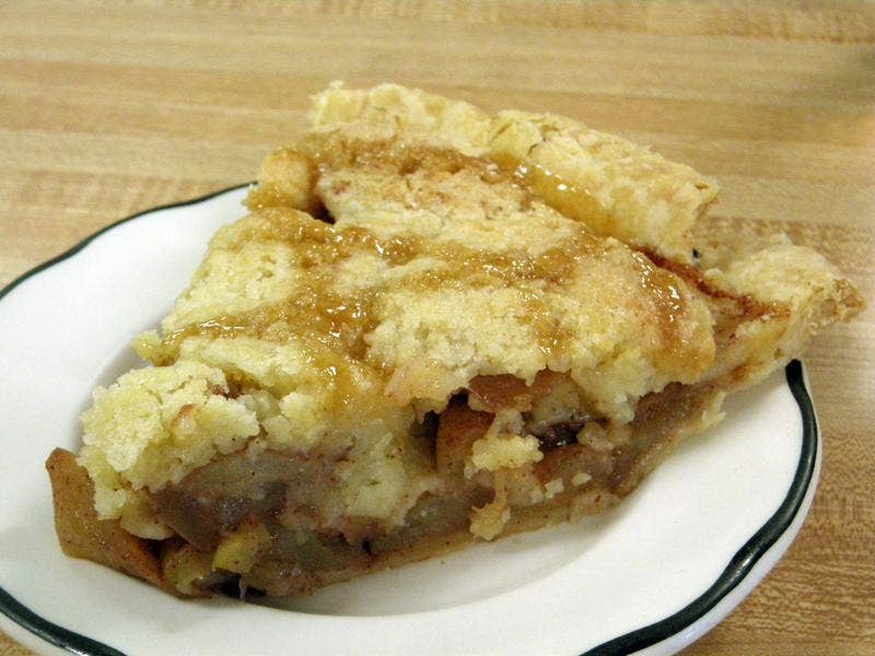 A slice of apple pie with a crumbly golden crust and caramelized topping sits on a white plate with a dark rim, highlighting why classic apple pies remain a timeless dessert favorite on any light wooden surface.
