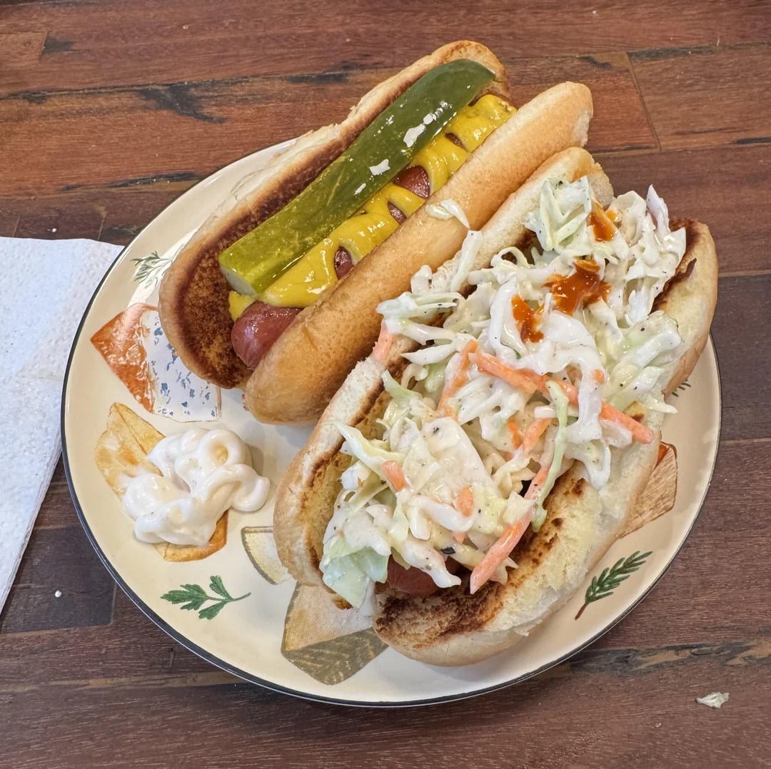 Two hot dogs on a plate: one with mustard and a pickle spear, the other topped with coleslaw and hot sauce. A dollop of mayonnaise and a potato wedge are also on the plate, all set on a wooden table.
