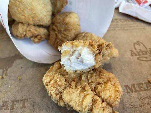 Close-up of three pieces of golden, crispy fried chicken tenders, one showing the white, juicy inside. The tenders are on a brown paper wrapper next to a white container.