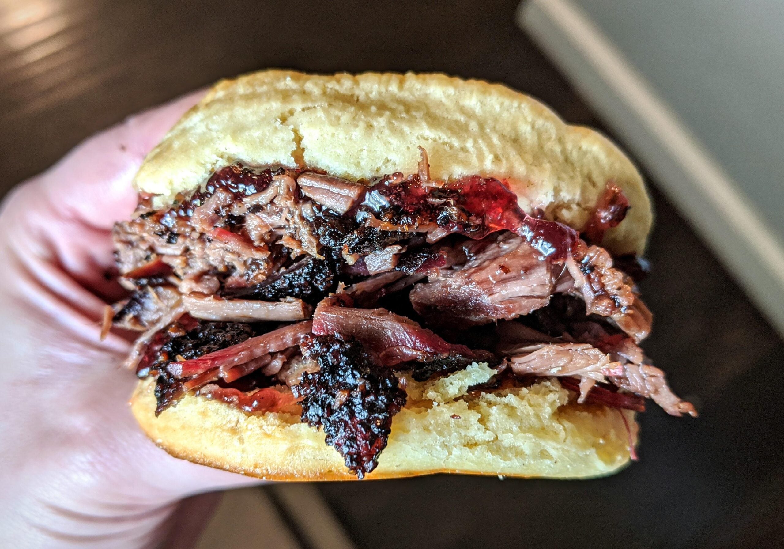 A hand holds a sandwich filled with shredded, juicy barbecue beef on a soft bun, with visible charred edges and sauce.
